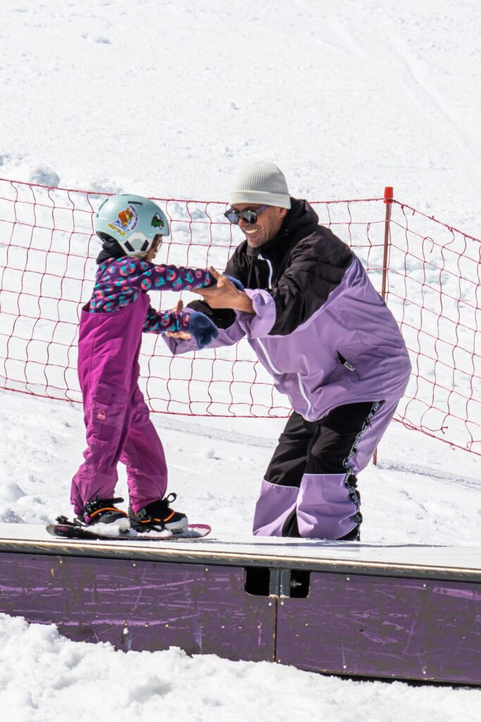 Enfant en équilibre sur un module de snowboard aidé par un moniteur – cours enfants aux 2 Alpes